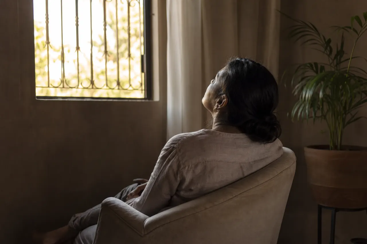 Una mujer sentada en un sillón mira hacia la ventana, pensativa